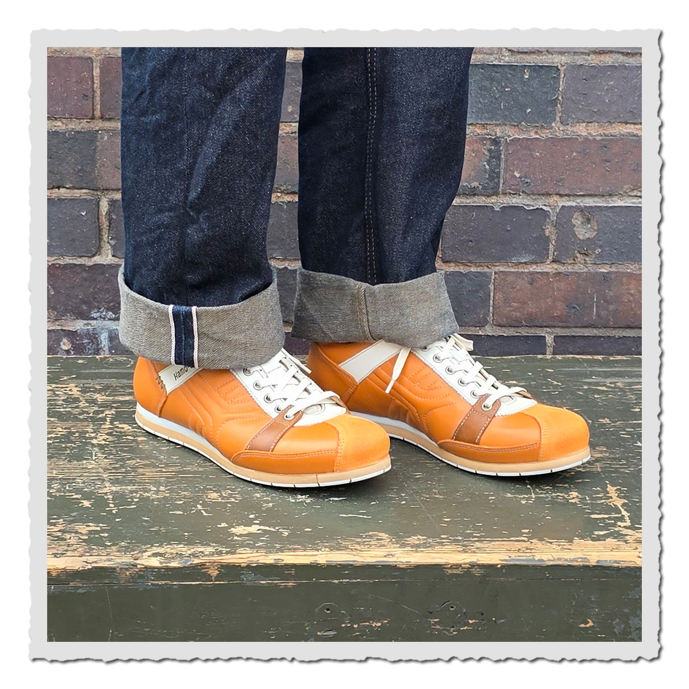 Orange Schnürschuhe an einer Person mit umgeschlagenen dunklen Jeans, auf grün lackierter Holzfläche vor Backsteinwand.